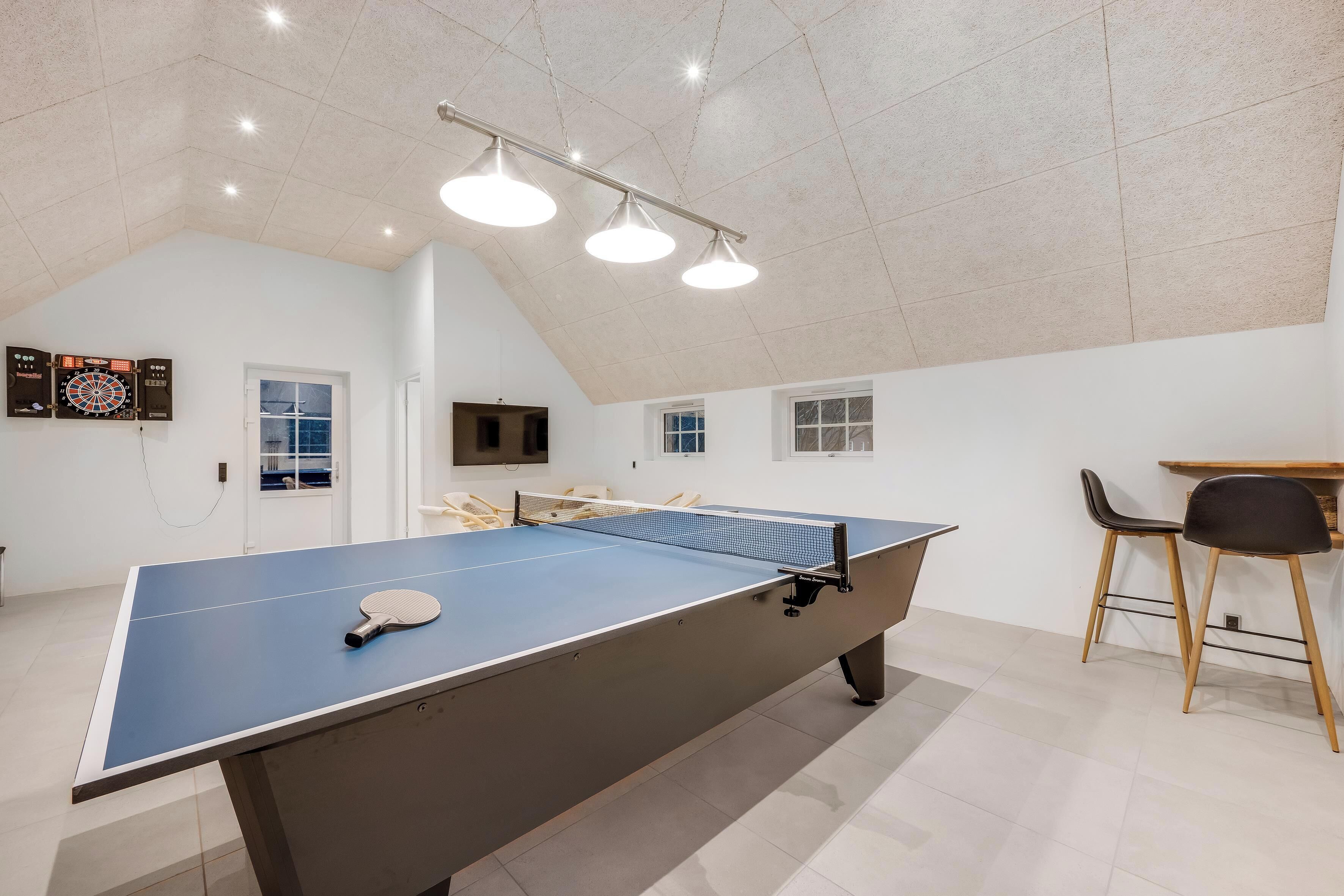 A ping pong table with a paddle, a dartboard, and a TV mounted on the wall.