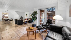 Ein großzügiger Wohnbereich mit Esstisch, Ledersofas, einem Couchtisch mit Obst und einem großen Fenster mit Aussicht.