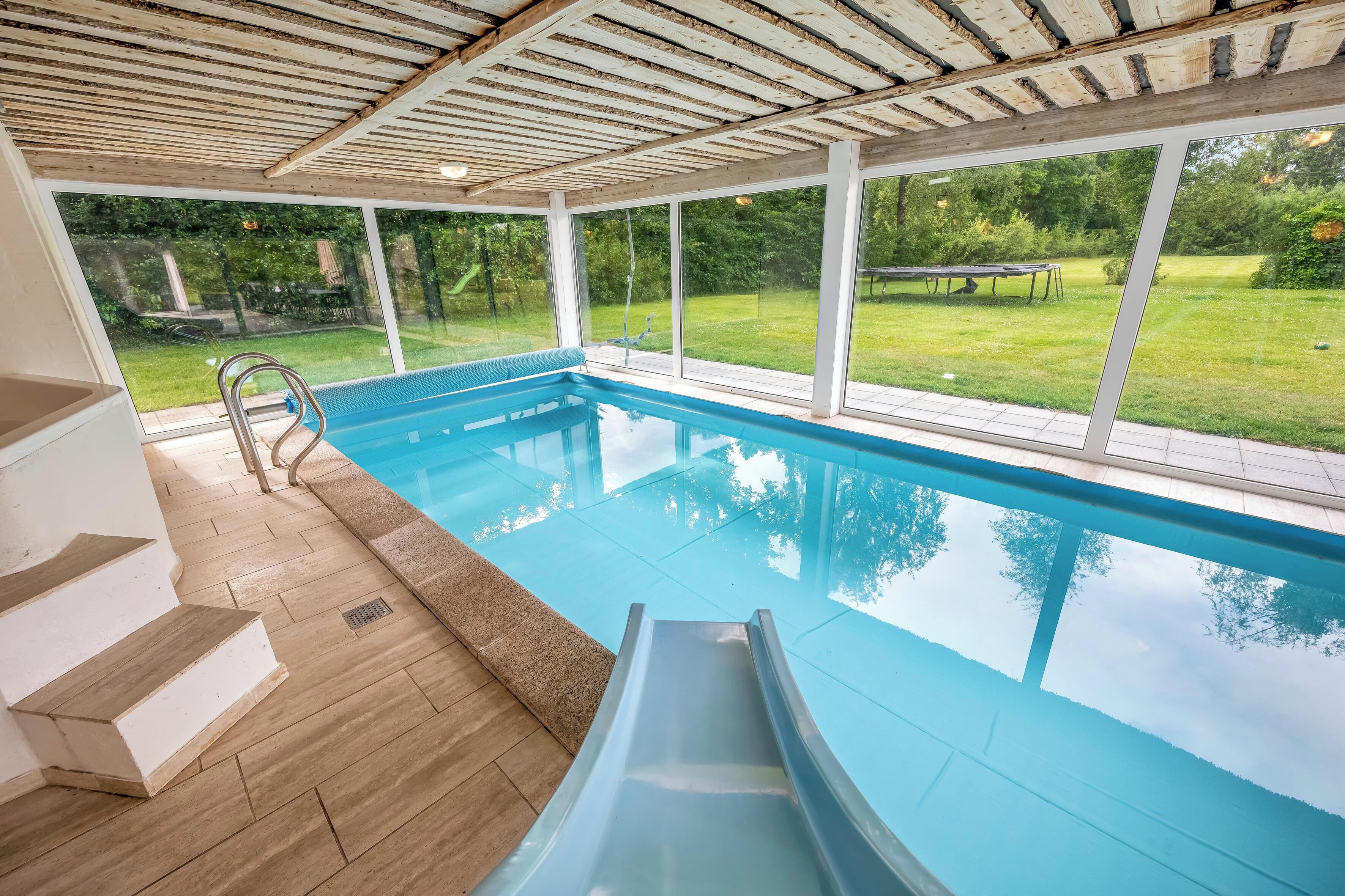 Una piscina interior con tobogán, escalones de madera y vistas a un césped.