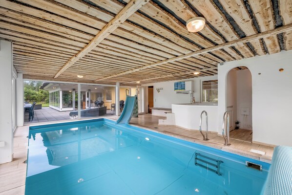 An indoor swimming pool with a wooden ceiling, a slide, and a view of the outdoors.