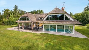 Ein modernes Haus mit großem, verglasten Pool, einem Strohdach und einem großzügigen Essbereich im Freien.