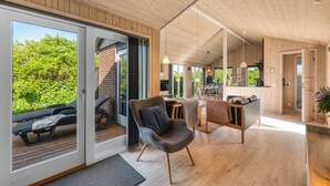 Een ruime kamer met een houten plafond, een schuifdeur naar een terras met ligbedden, een eethoek met tafel en stoelen, en een keuken met een aanrecht en hangende lampen.