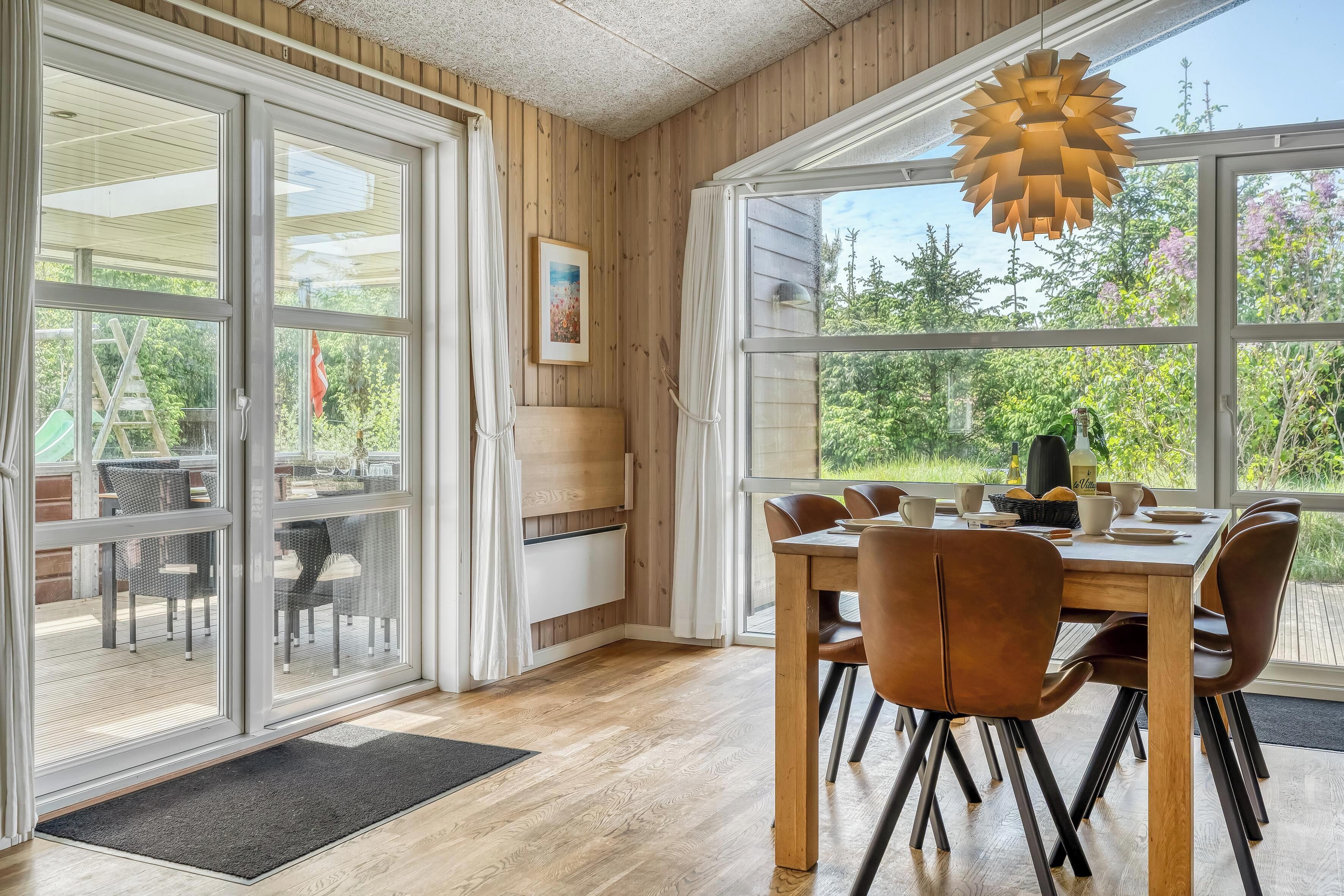 A dining area with a wooden table and chairs, a large window with a view of greenery, and a ceiling light fixture.