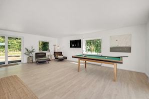 A modern living room with a pool table, comfortable seating, a flat-screen TV, and large windows.