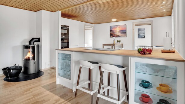 Een moderne keuken met een houtkachel, witte barkrukken en een glazen kast met kopjes.