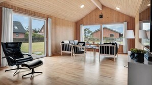 Een ruime kamer met houten vloer, een zwarte leren fauteuil, een witte bank, een kleine tafel en grote ramen met uitzicht op buiten.