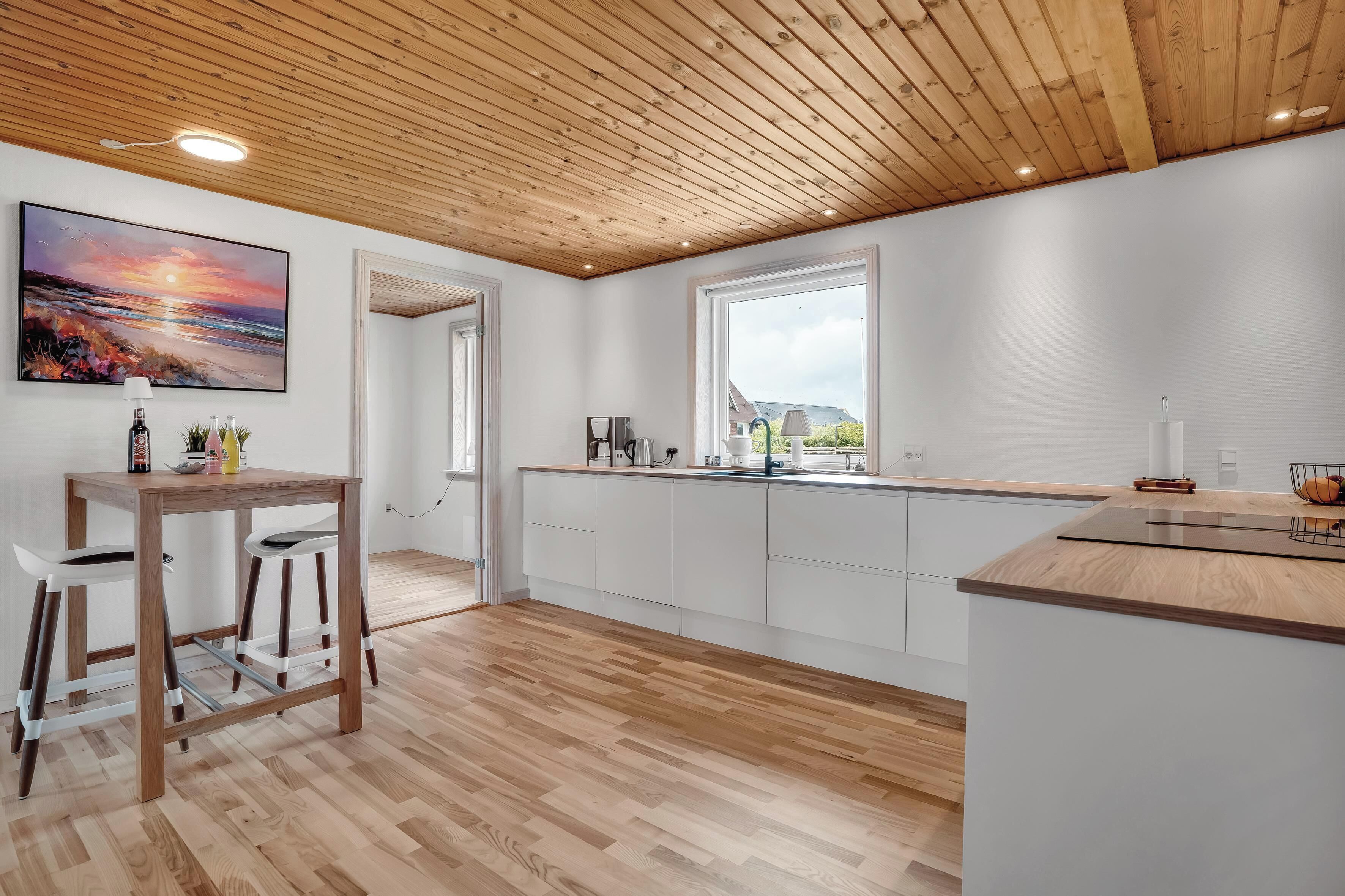 Une cuisine moderne avec un plancher en bois, des meubles blancs, un plafond en bois et une grande fenêtre.