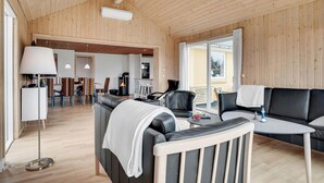 Een moderne woonkamer met een houten plafond, een zwarte leren bank, een witte fauteuil, een eethoek met een tafel en stoelen, en een keuken op de achtergrond.