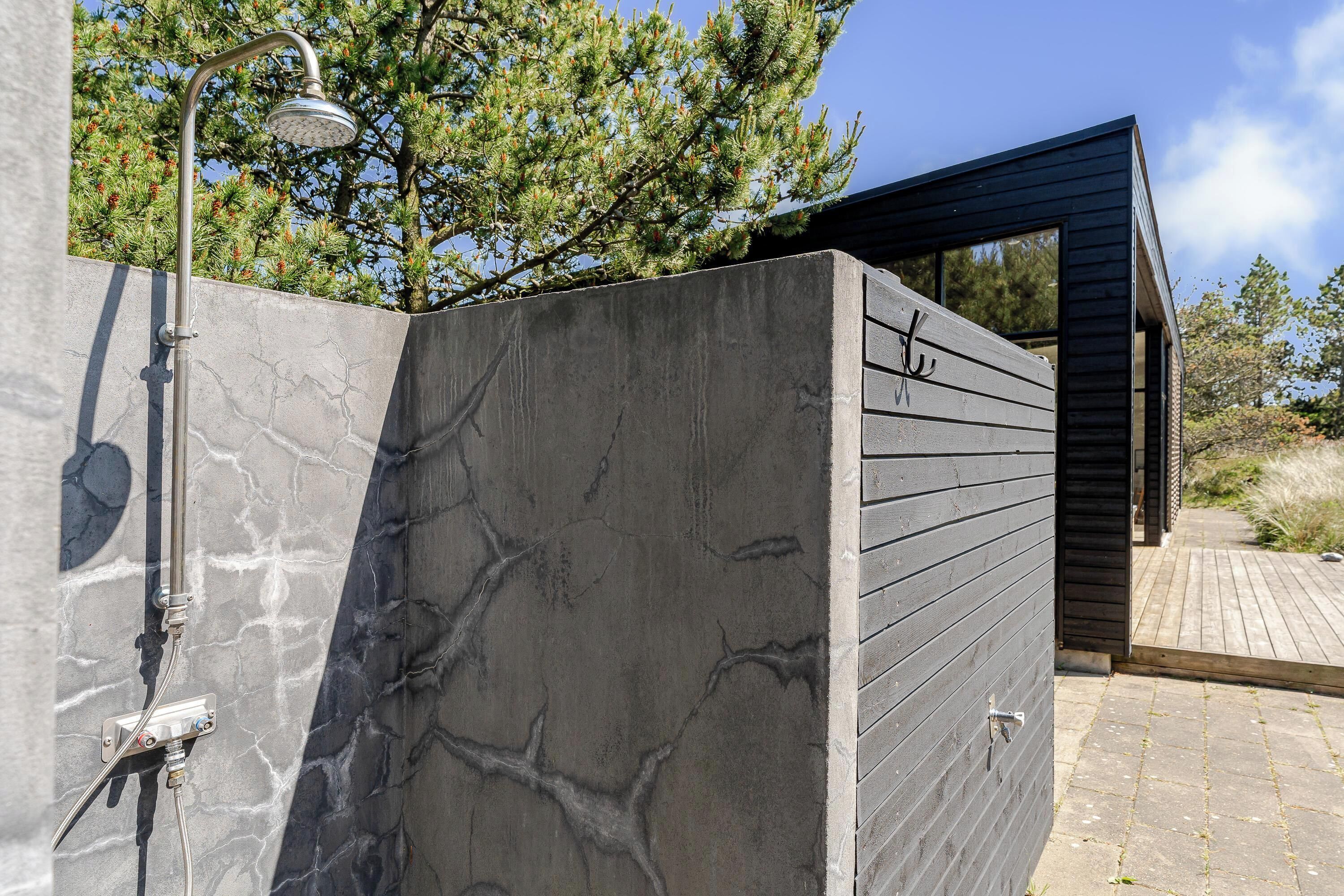 Une douche extérieure moderne dotée d’un pommeau de douche en métal imitant la pluie, d’un mur en béton au motif évoquant le marbre et d’une terrasse en bois.
