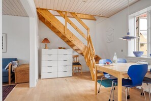 Un interior moderno con una escalera de madera, una zona de comedor con sillas azules, un armario blanco y un ventanal con luz natural.