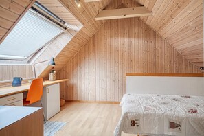 Une petite chambre mansardée avec un plafond en pente, une lucarne, un bureau avec une chaise, un lit avec une courtepointe et une petite armoire.
