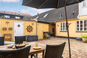Une terrasse extérieure avec des meubles en osier, une table avec des assiettes et un panier, une porte ornée d’un motif de roue de navire, et un bâtiment jaune avec un toit en tuiles.
