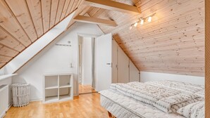 A bedroom with a sloped ceiling, wooden beams, a bed with patterned bedding, a white wardrobe, and a small white cabinet.