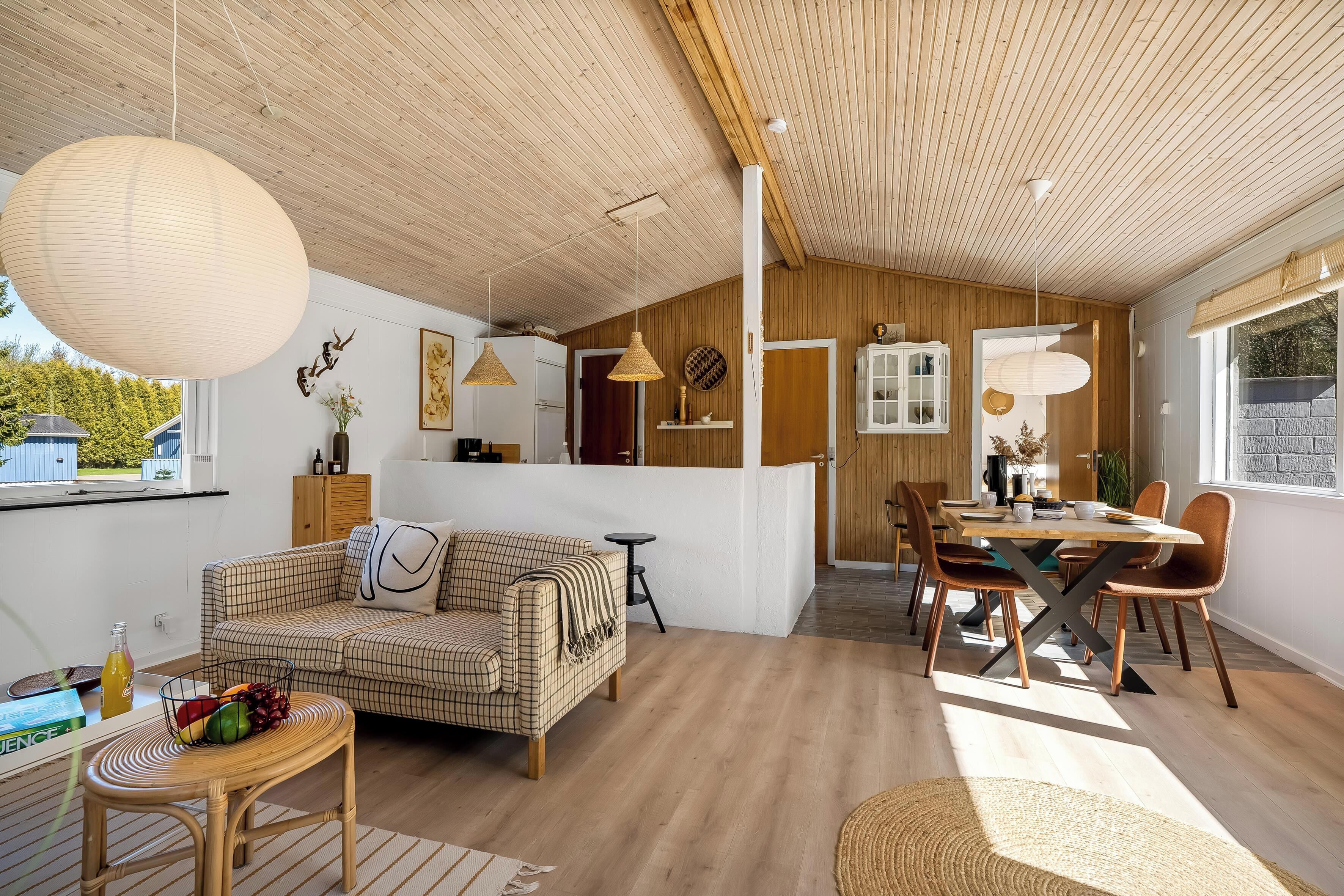Et moderne interiør med træloft, spiseområde med træbord og stole, sofa og et fletbord med frugt.
