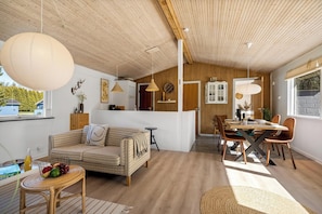Un interior moderno con techo de madera, una zona de comedor con mesa y sillas de madera, un sofá y una mesa de mimbre con fruta.