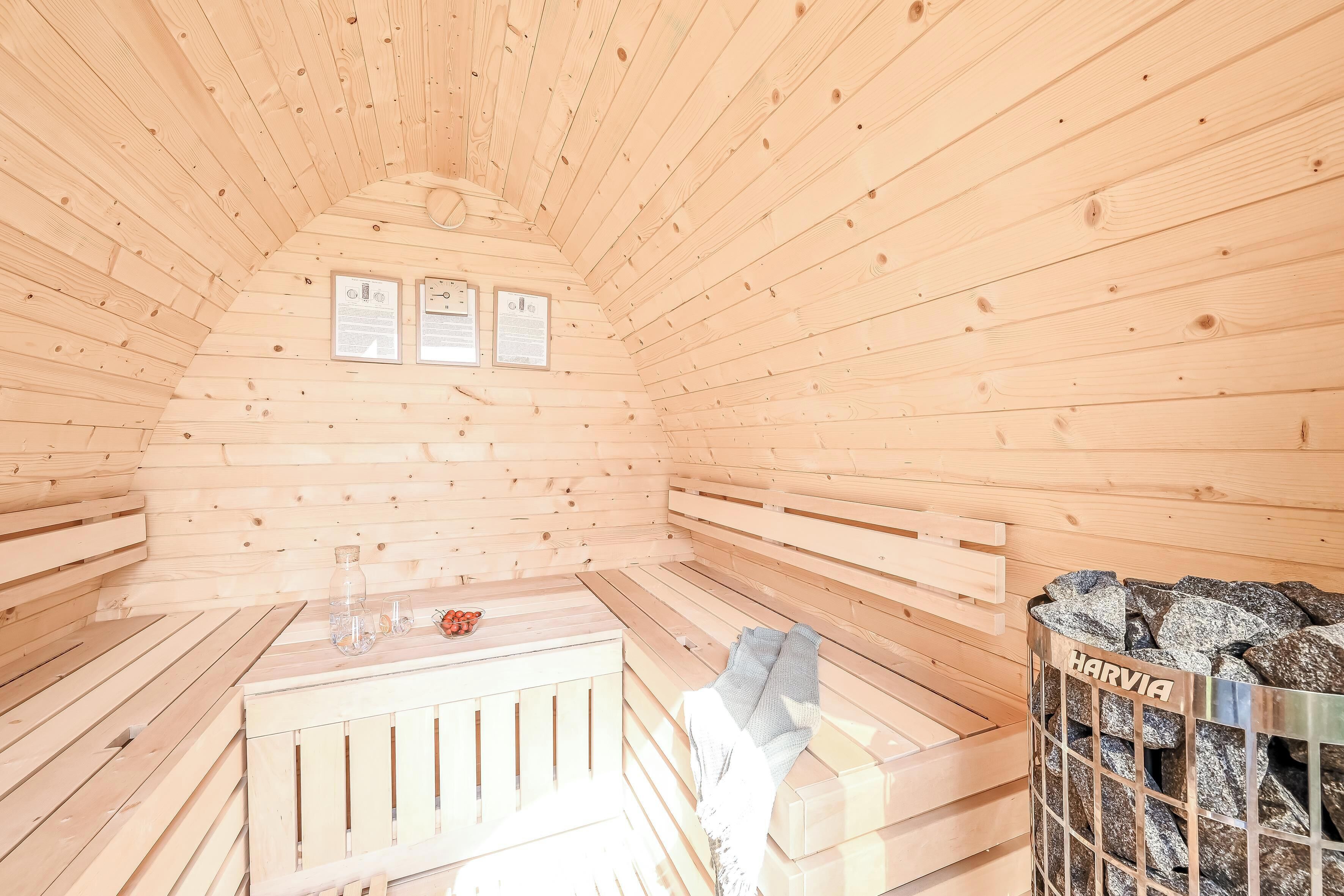 Een houten sauna met banken, een bank met een handdoek, een glazen vat en een metalen mandje met de tekst 'HARVIA'.
