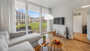 Un soggiorno moderno con un divano, un tavolino rotondo con una pianta e della frutta, una televisione e un'ampia finestra vista un edificio e un prato.