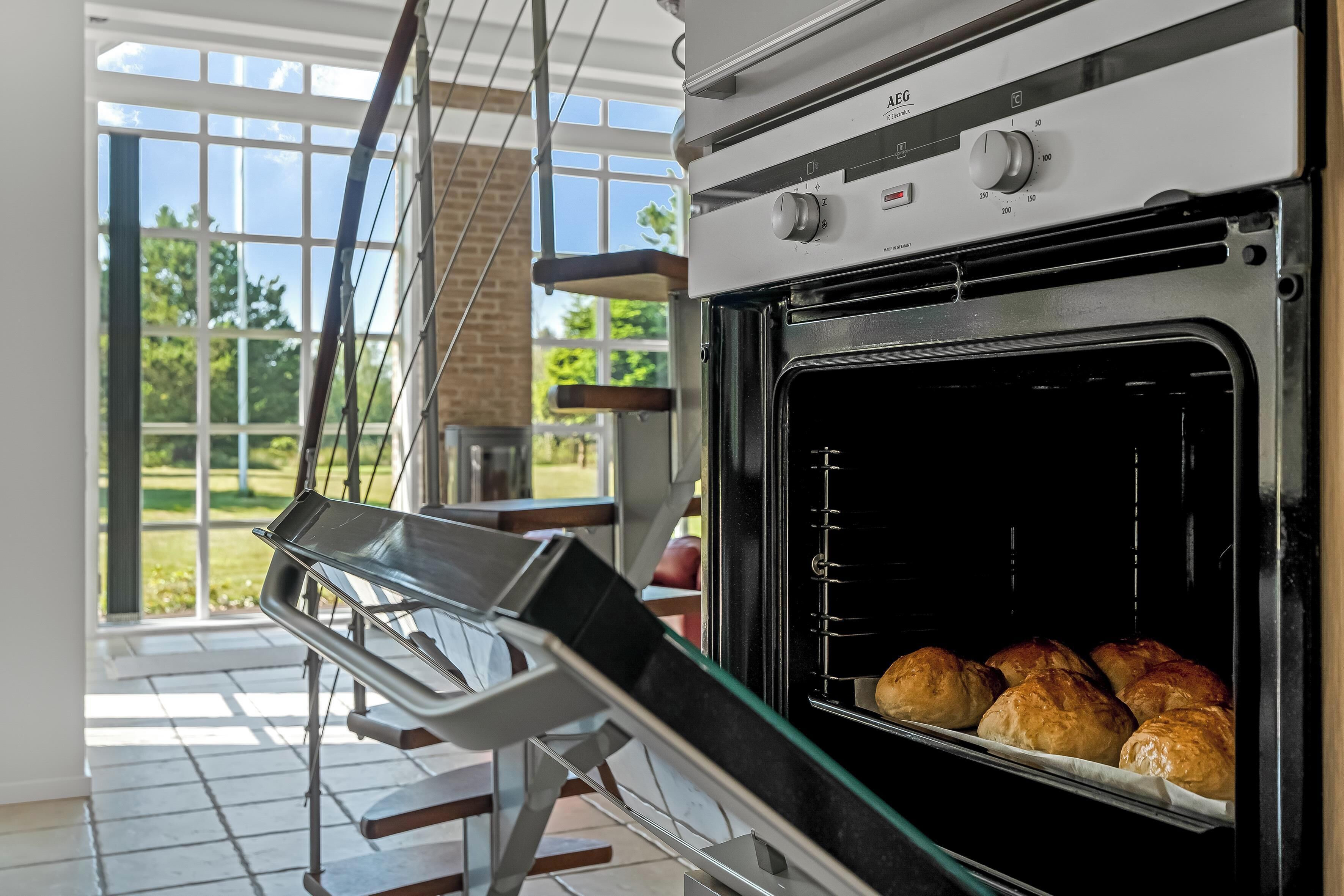 An oven with a tray of bread inside, a window with a view of greenery, and a hanging rack.