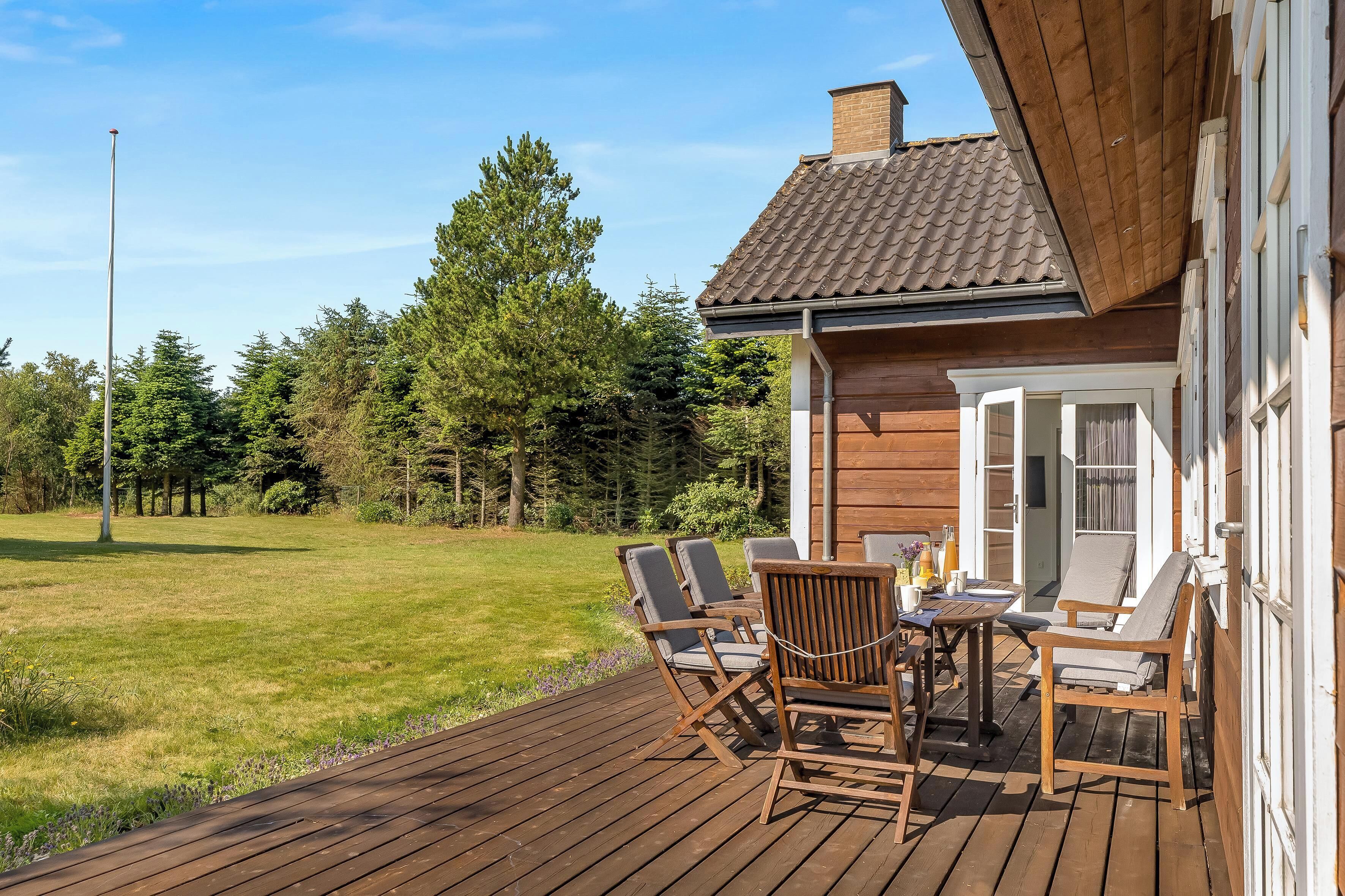 Um deck de madeira com mobiliário externo, vista desobstruída para uma floresta e um edifício com telhado de azulejos.