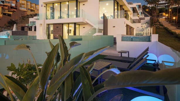 Un edificio moderno de varios pisos con balcones de vidrio, una zona de piscina y mobiliario exterior.