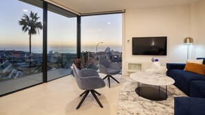 Un salón moderno con un televisor grande, dos sillones grises, un sofá azul y vistas a la ciudad a través de amplias ventanas.