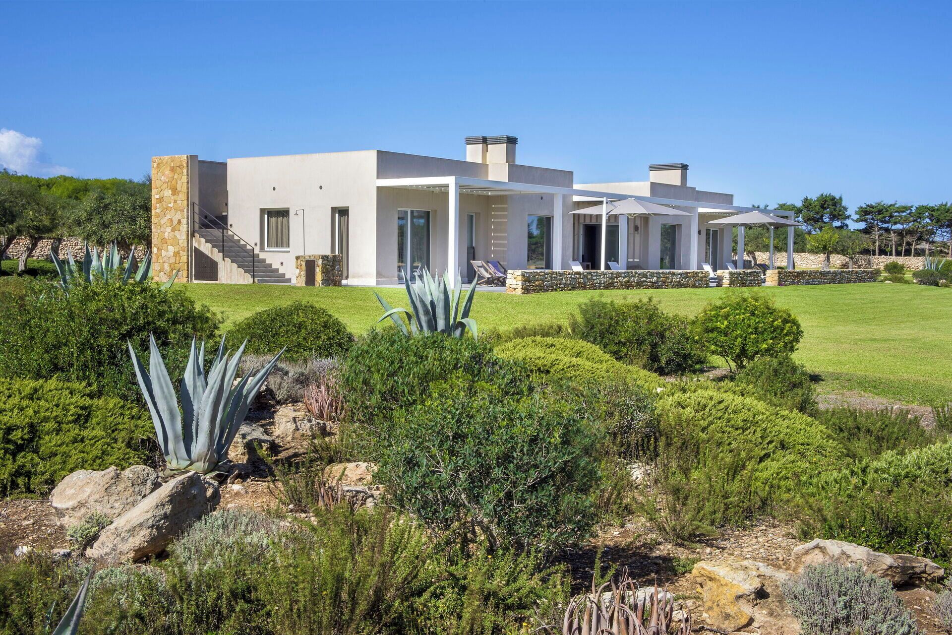 Une maison moderne à deux étages, dotée d’une façade en pierre, d’un patio couvert avec des parasols et d’un jardin bien entretenu où poussent diverses plantes et arbustes.