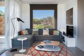 Ein modernes Wohnzimmer mit einer Eckcouch, einem Flachbildfernseher, einer Stehlampe und einem großen Fenster mit Blick auf Bäume.