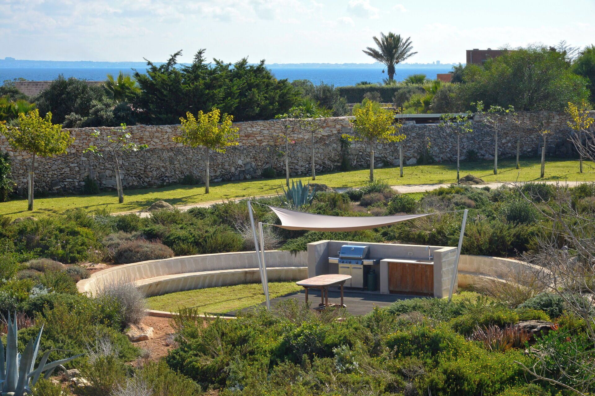 Un área exterior ajardinada con una zona de barbacoa, muros de piedra y vistas al mar.