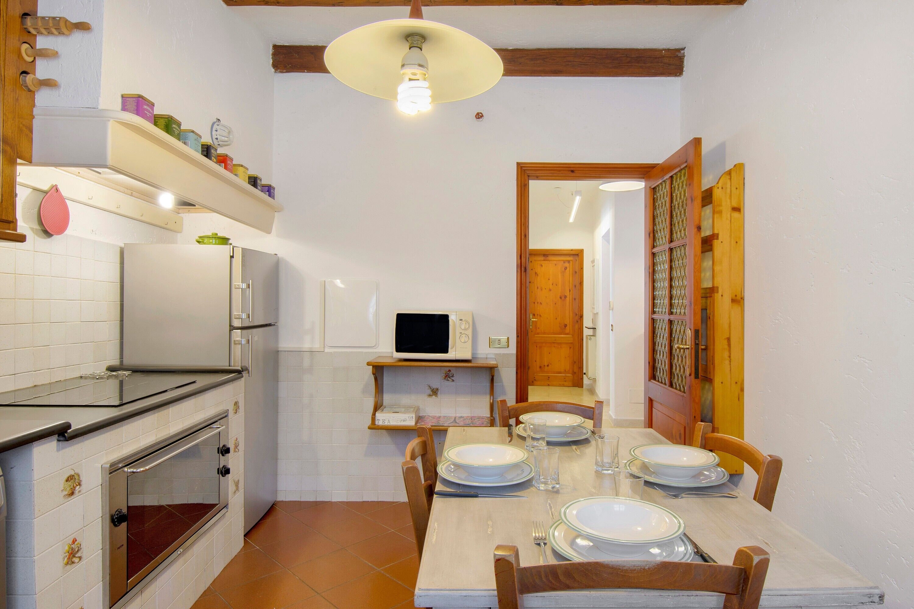 A kitchen with a microwave, oven, and shelves with jars. There is a dining table with plates and chairs.