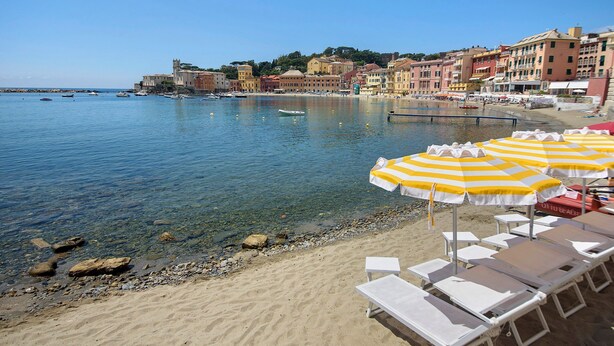 A beach with clear water, white lounge chairs, and yellow and white striped umbrellas.