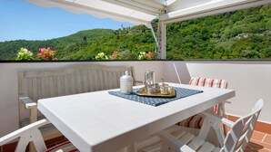 A balcony with a white table and chairs, a metal tray with cups, a white container, and a view of a green hillside.
