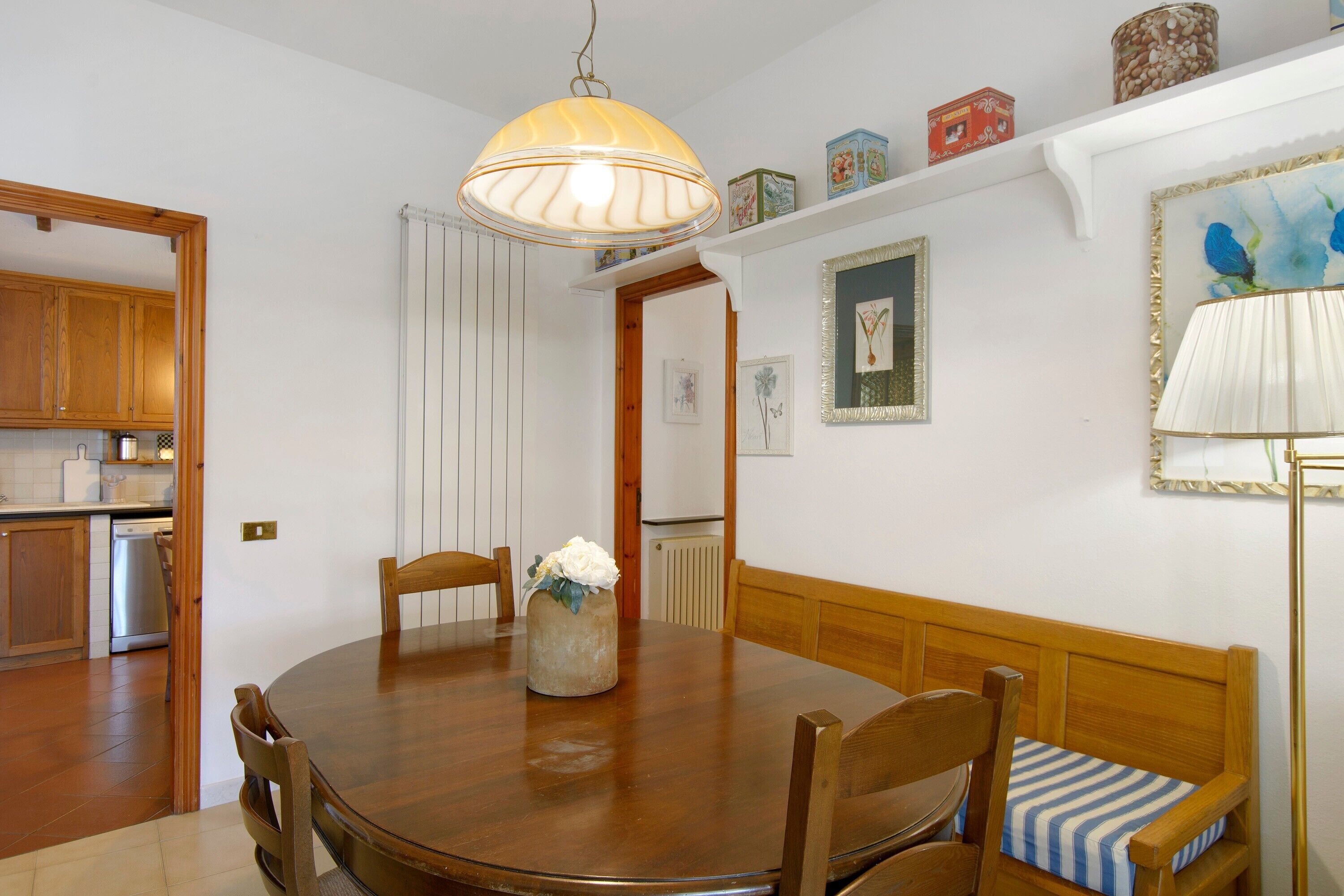 A dining area with a wooden table, chairs, a vase with flowers, a hanging lamp, and a view into the kitchen.