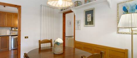 A dining area with a wooden table, chairs, a vase with flowers, a hanging lamp, and a view into the kitchen.