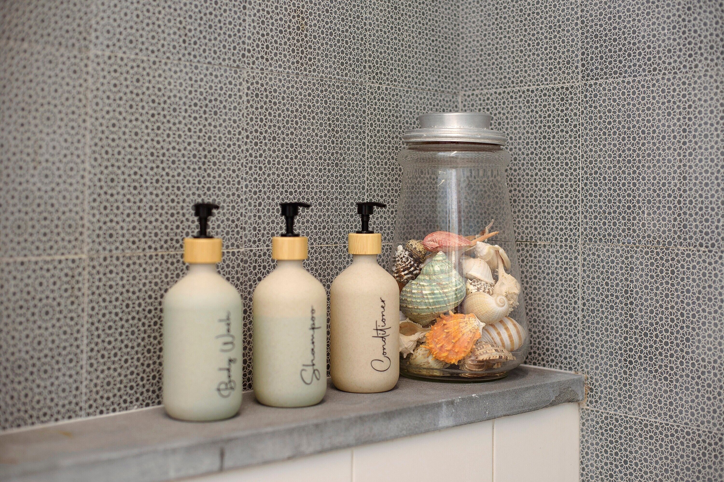 Drei Flaschen Shampoo und Conditioner auf einer Arbeitsplatte mit einem Glasbehälter voller Meeresmuscheln.
