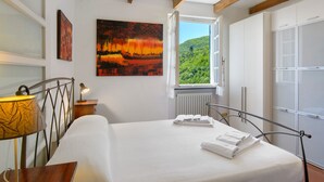 A bedroom with a bed, a window with a view of greenery, a painting on the wall, and a lamp.