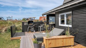 Une terrasse en bois avec des meubles d’extérieur, un barbecue et une fenêtre donnant sur un espace verdoyant.