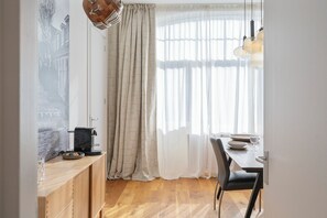 Un coin repas avec une table en bois, des chaises, une machine à café et une vue dégagée à travers des rideaux légers.