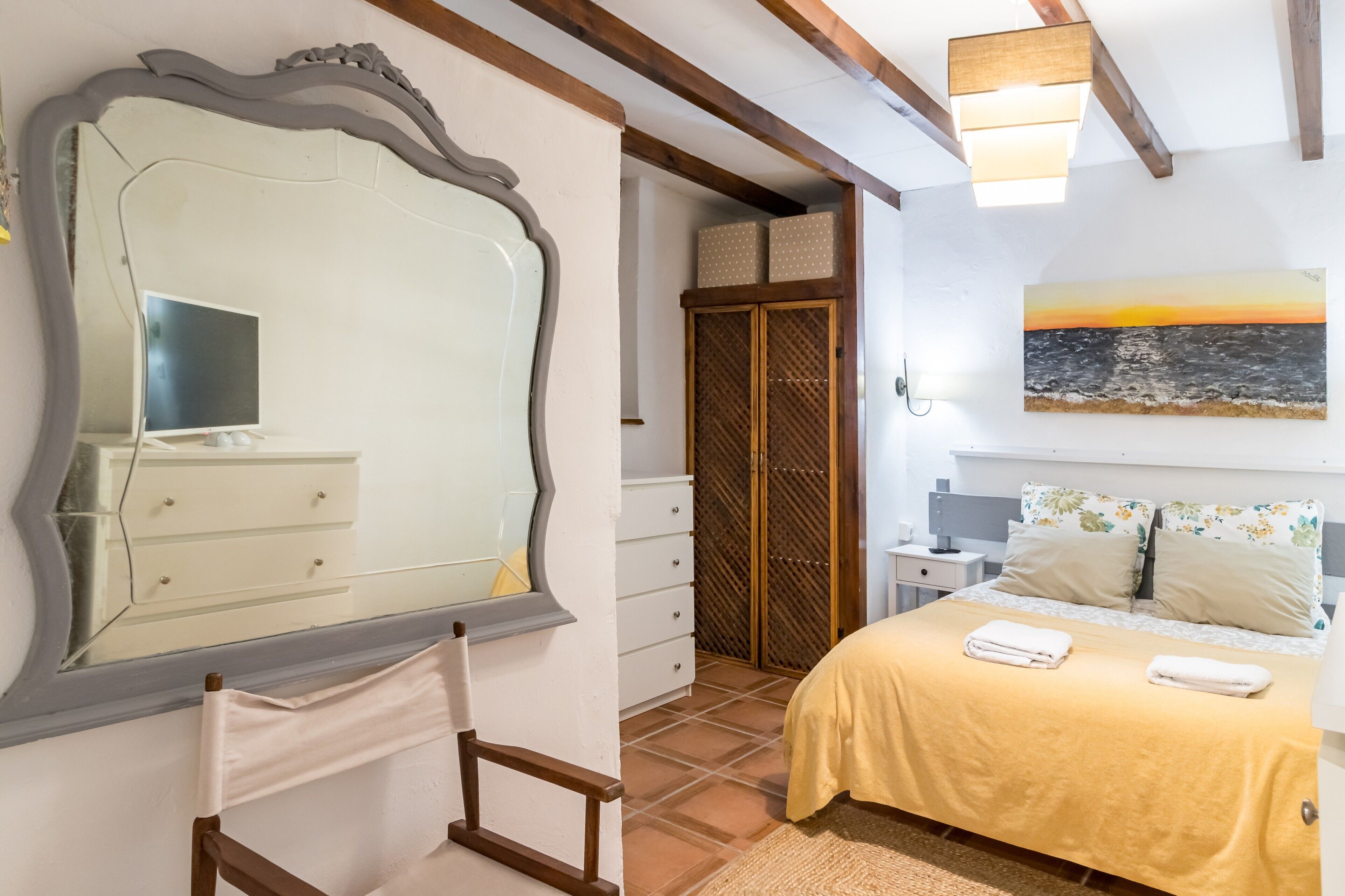 Un dormitorio con un espejo grande, una cama con colcha amarilla, un armario de madera y un cuadro de una puesta de sol.