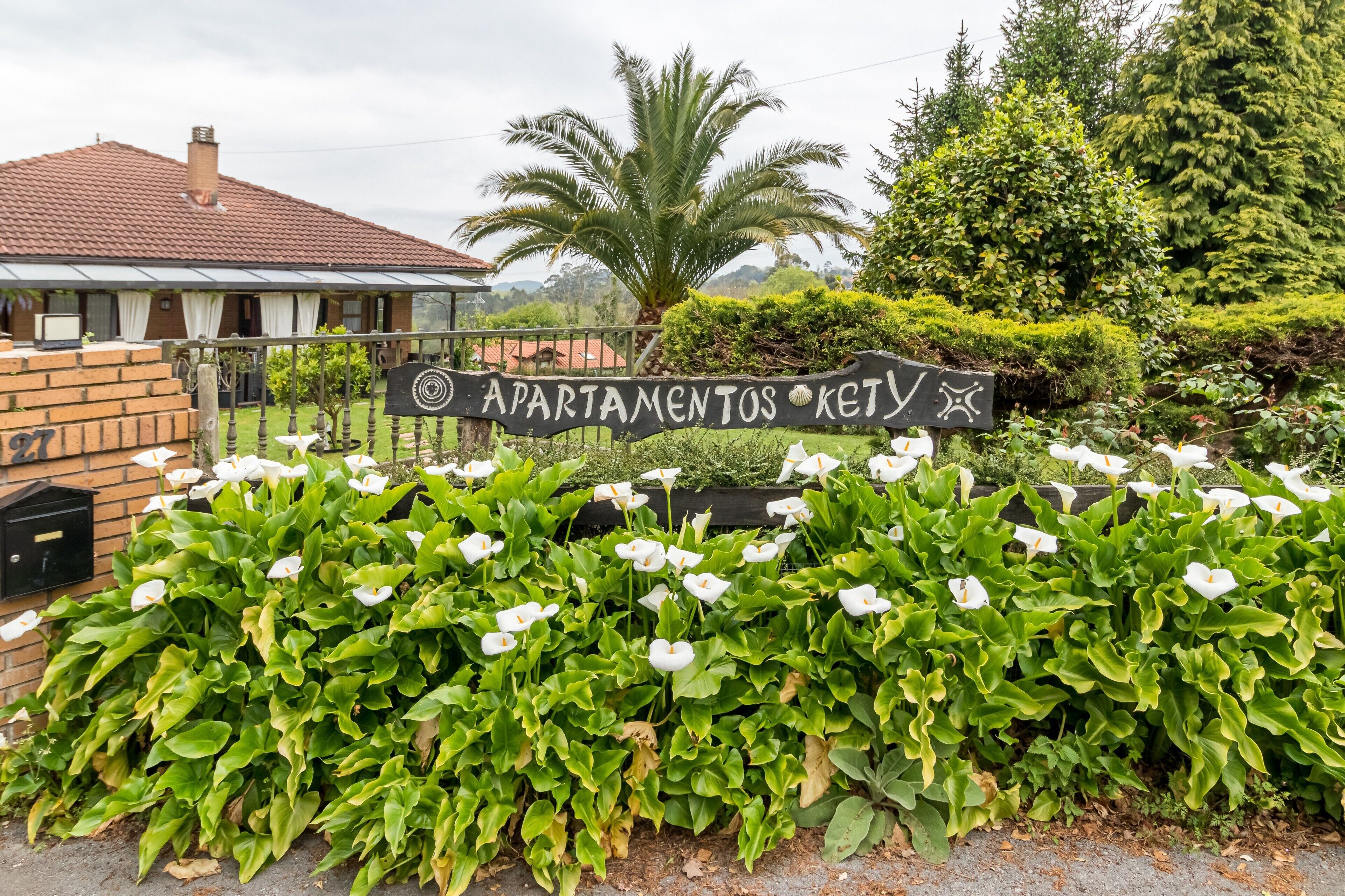 Un letrero que dice 'Apartamentos KETY', con un edificio de ladrillo y abundante vegetación al fondo.