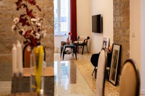 A room with a marble floor, a dining area with a table and chairs, a TV mounted on the wall, and a window with red curtains. - ELITE COLLECTION- MAISON NOIR (Naples)