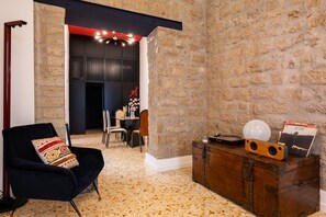 A room with a stone wall, a black armchair, a wooden cabinet with decorative items, and a dining area in the background. - ELITE COLLECTION- MAISON NOIR (Naples)