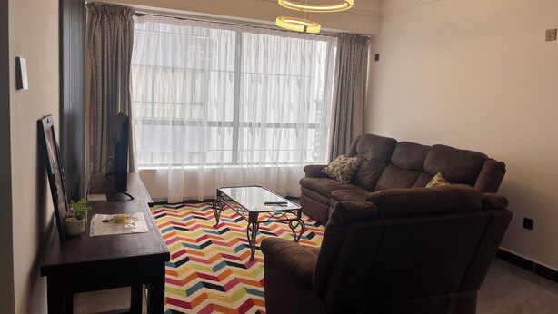 Uma sala de estar com um sofá, uma mesa de centro de vidro, uma televisão em um móvel e um tapete colorido.