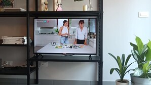 Un salon avec un téléviseur diffusant une émission de cuisine, une étagère noire contenant des livres et des objets de décoration, et une plante en pot.
