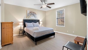 Une chambre à coucher avec un lit, un ventilateur de plafond, une fenêtre, une commode en bois et une chaise.