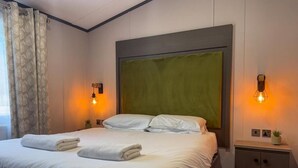 A neatly made bed with white linens and towels, a green headboard, two wall-mounted light fixtures, and a small bedside table with a plant. - Beck Lodge with Hot tub (Allerthorpe)