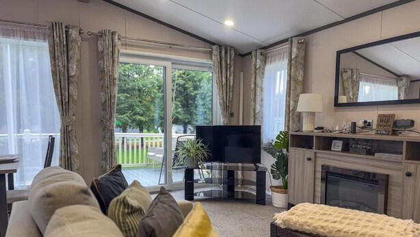 A living room with a sofa, a television, a fireplace, and a balcony with a view of greenery. - Beck Lodge with Hot tub (Allerthorpe)