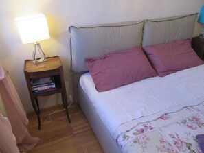 Un letto rifatto con cura, con una coperta a fiori e due cuscini, una lampada da comodino e un piccolo comodino di legno con dei libri.