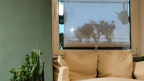 Ein Zimmer mit Holzboden, einem Fenster mit Blick auf Bäume und eine Straßenlaterne, einer Topfpflanze und einem beigefarbenen Sofa.
