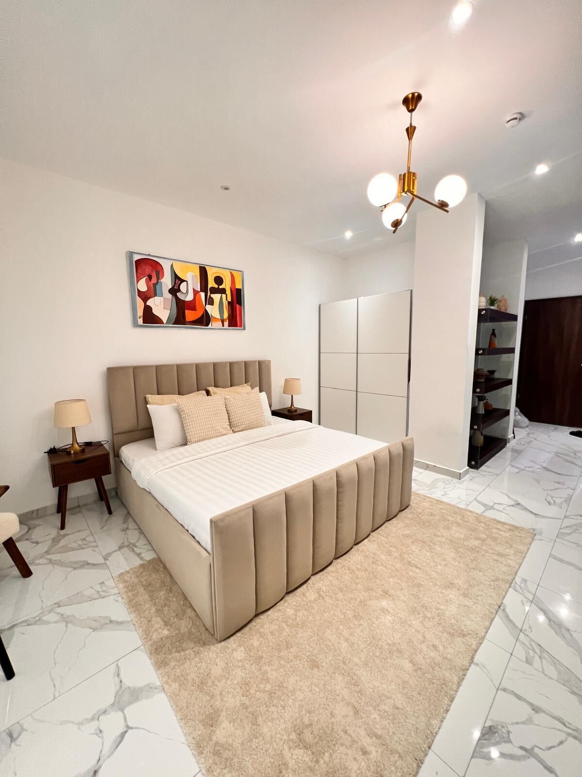 Une chambre à coucher comprenant un lit, des tables de chevet, une lampe, un luminaire au plafond, une œuvre d’art accrochée au mur et une armoire.