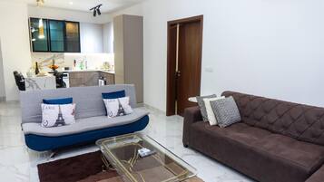 A living room with a sofa, a brown couch, a glass coffee table, and a kitchen in the background.